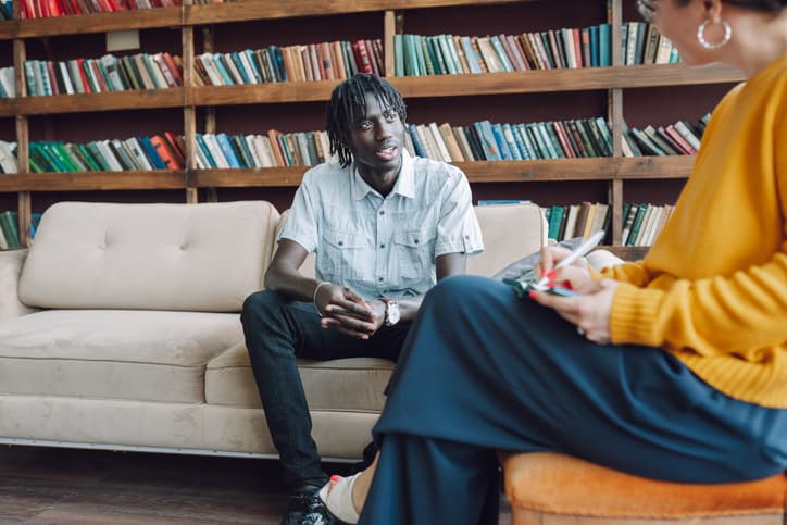 Young man in therapy. A young man sit on a sofa in a therapist office against female counselor