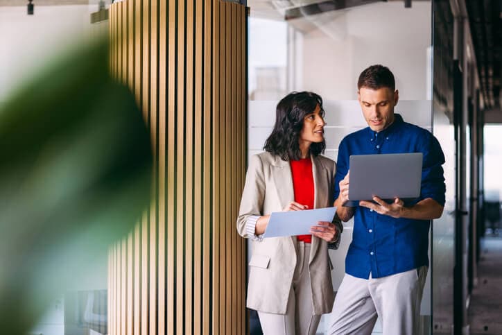 Two professionals discussing strategies using a laptop and documents in a modern office space.