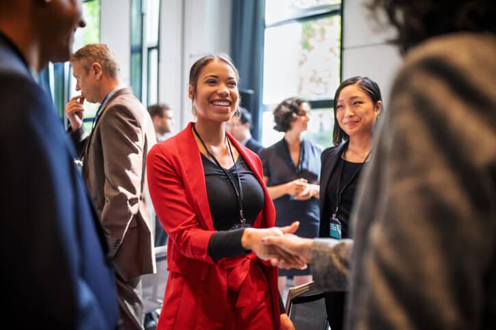 Confident Black businesswoman shaking hands with another conference attendee while standing in the corridor of an auditorium at a networking event.