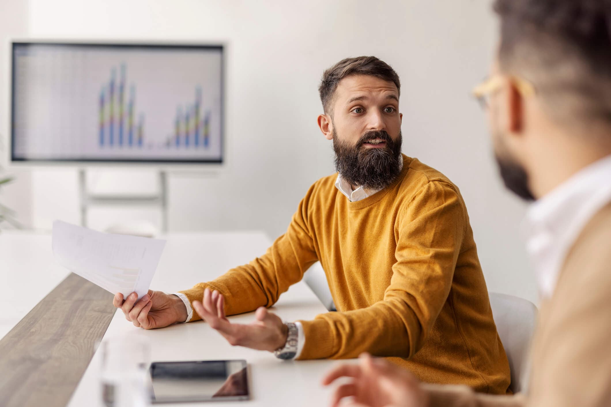 A bearded man wearing a mustard-yellow sweater discusses product analytics with a colleague in a modern office. A screen displaying charts is in the background.