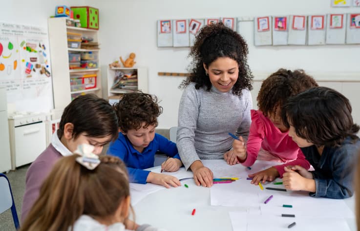 Focused school kids coloring with crayons while cheerful teacher supervises.