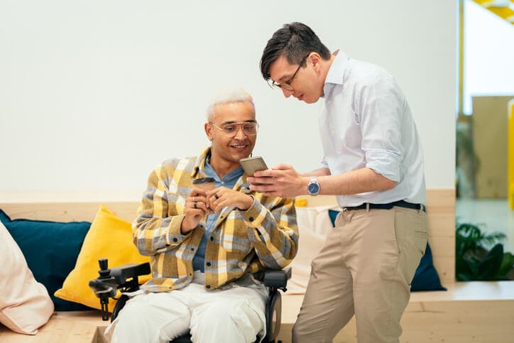 Male business leader sitting in a wheelchair looking at a colleague's smartphone at work.