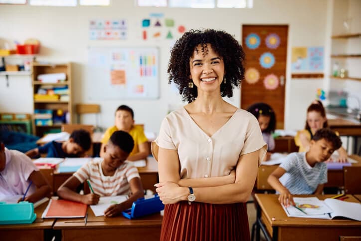 Young female teacher with curly hair in classroom, smiling at camera with arms crossed.