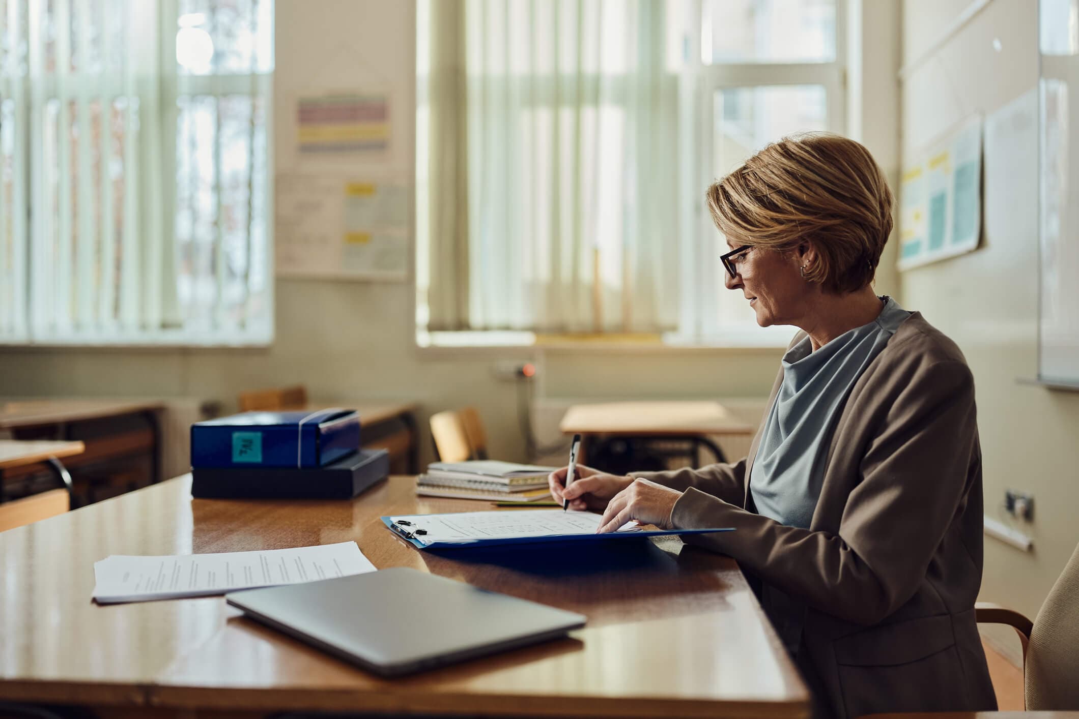 Senior female teacher grading exam papers in a high school classroom.