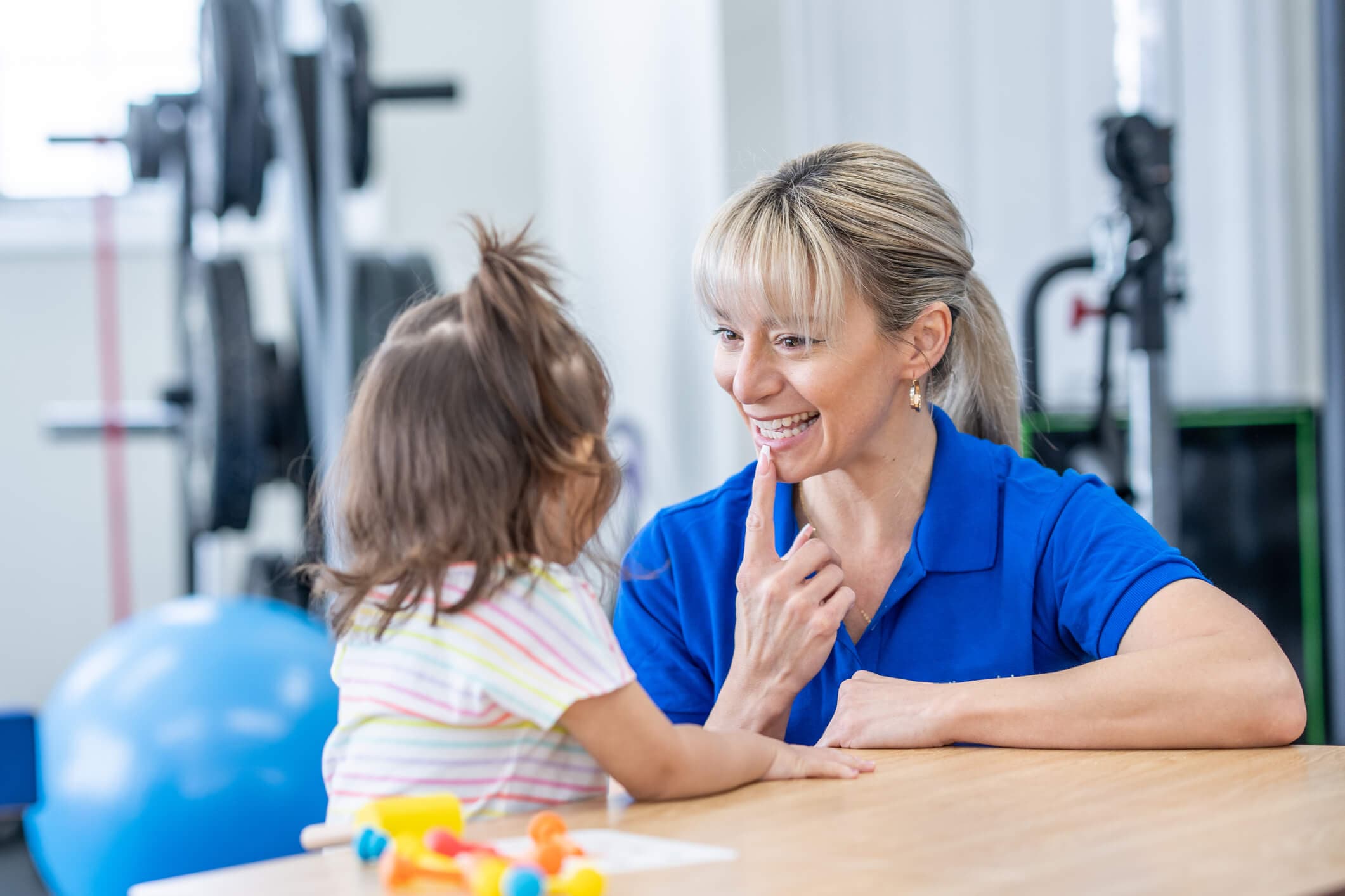 A female speech therapist sits with a little girl at a table as they work on her speech together. The therapist is pointing to her mouth to help show the girl how to form words.