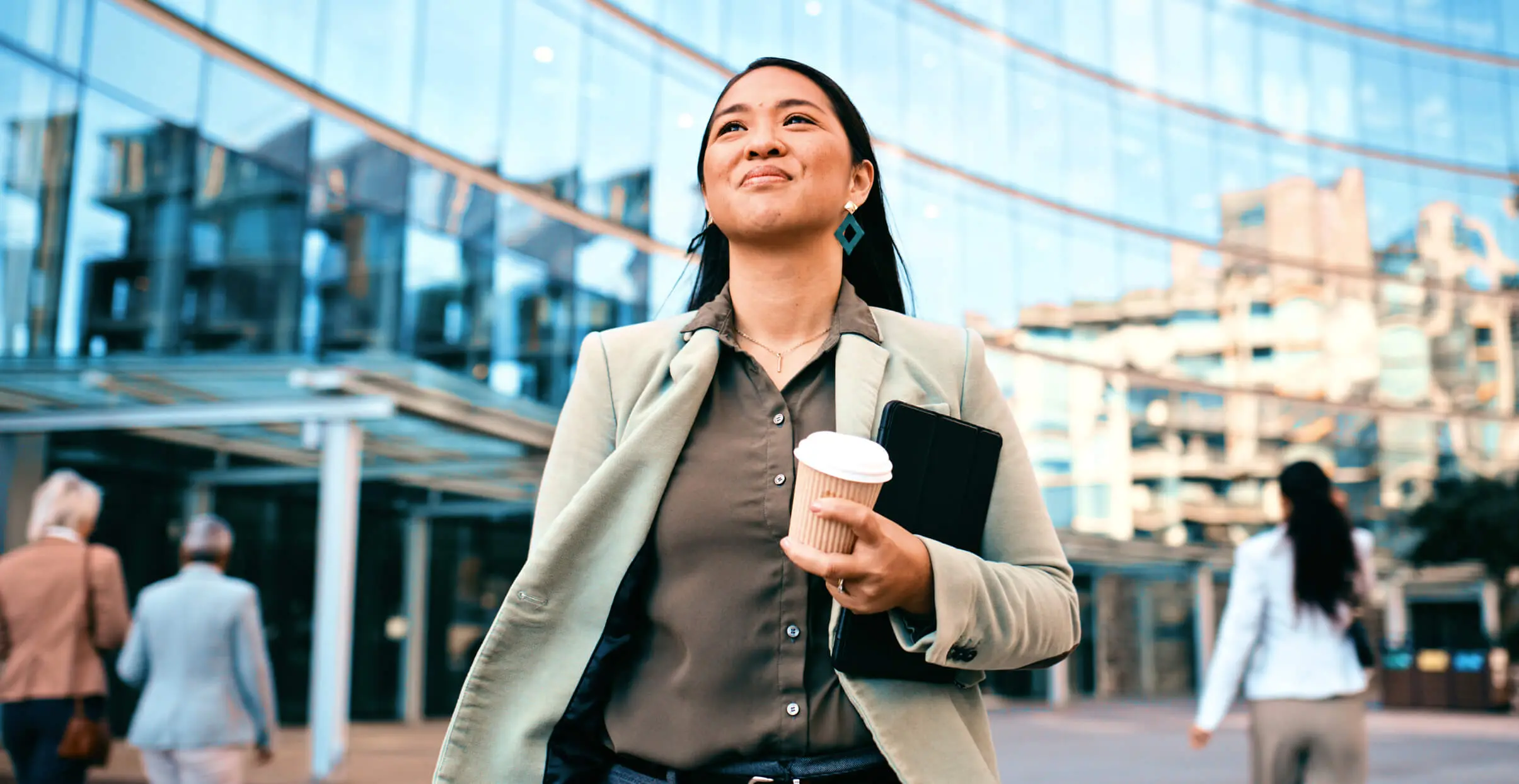 Smiling businesswoman holding coffee and documents while walking outside a modern office building.